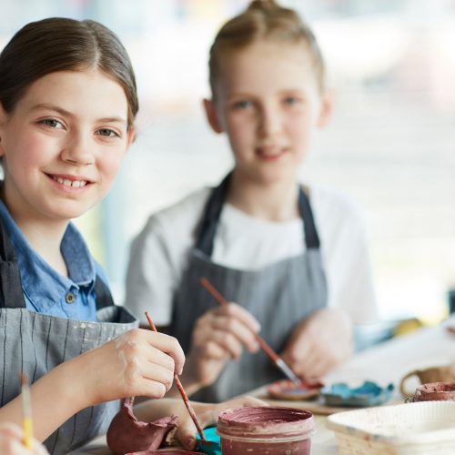 Little schoolgirl with paintbrush and her classmate sitting by table at lesson and creating clay items