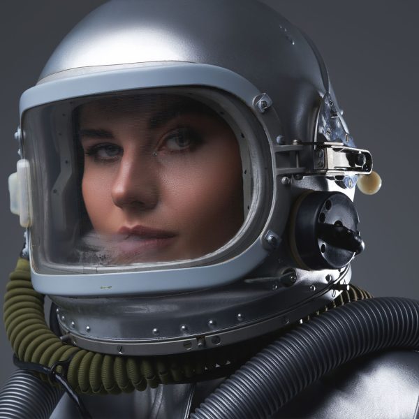 Studio headshot of young spacewoman with helmet against gray background.