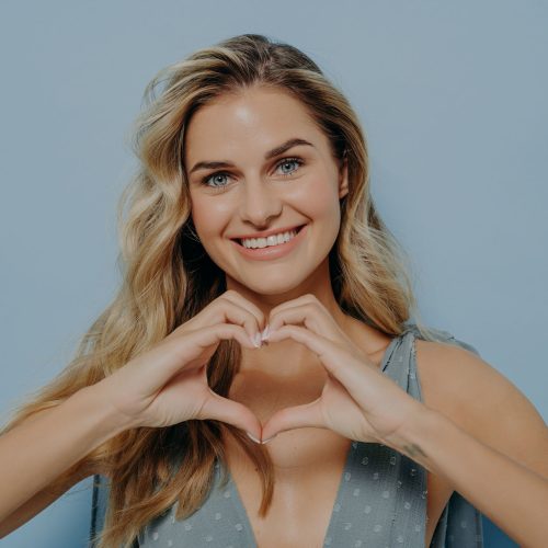 Image of lovely blonde woman in blue dress admitting in her love with use of heart gesture she made out of her hands, holding it in front while standing isolated next to blue background